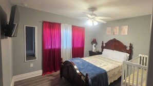 Quarto com cama, mesa de cabeceira, berço, ventilador de teto e janela com cortinas.