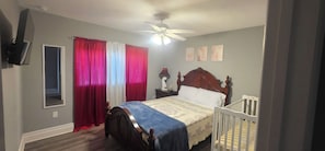 A bedroom with a bed, a nightstand, a crib, a ceiling fan, and a window with curtains. - Vrbo Property (Tampa)