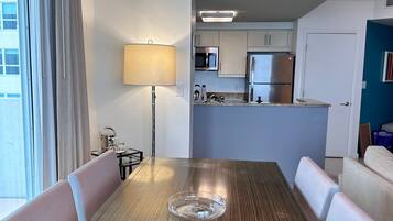 A dining area with a wooden table, white upholstered chairs, a lamp, and a view of a kitchen with modern appliances.