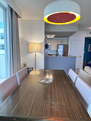 Un comedor con una mesa de madera, sillas blancas con tapicería, una lámpara y vistas a una cocina con electrodomésticos modernos.