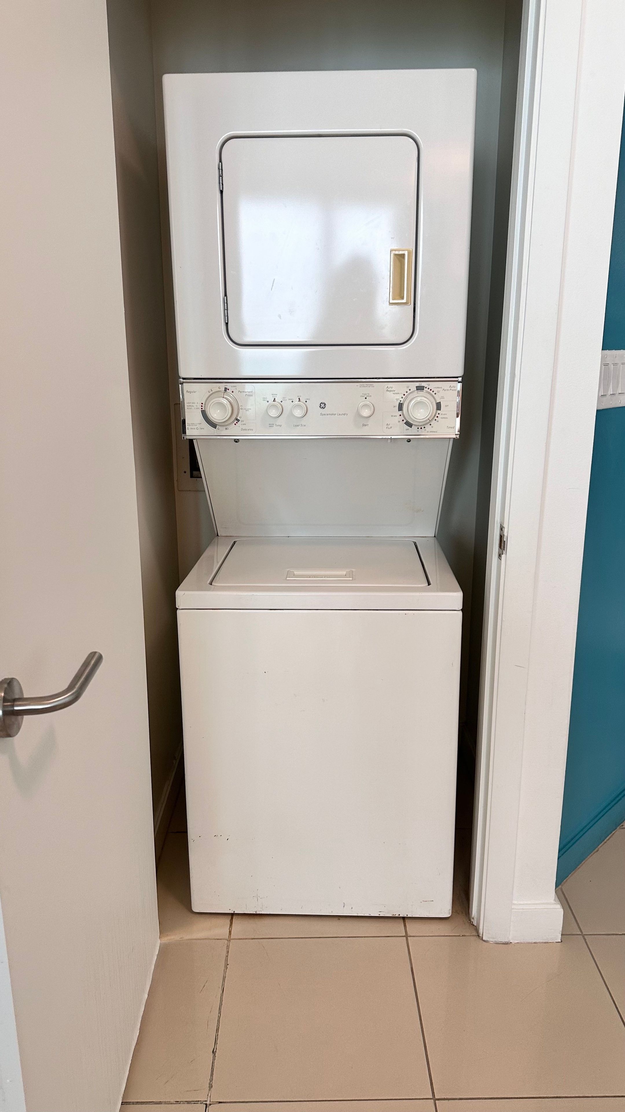 A compact, white laundry machine with a control panel, located in a small room with a tiled floor and a door on the left.