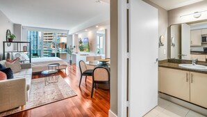 Habitación de hotel moderna con cama, zona de estar, mesa de comedor y baño con lavamanos y espejo.