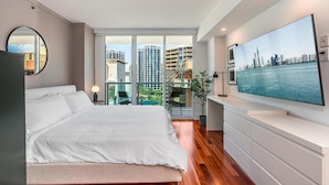 Un dormitorio moderno con una cama grande, un televisor sobre un mueble blanco y un balcón con vista a la ciudad.
