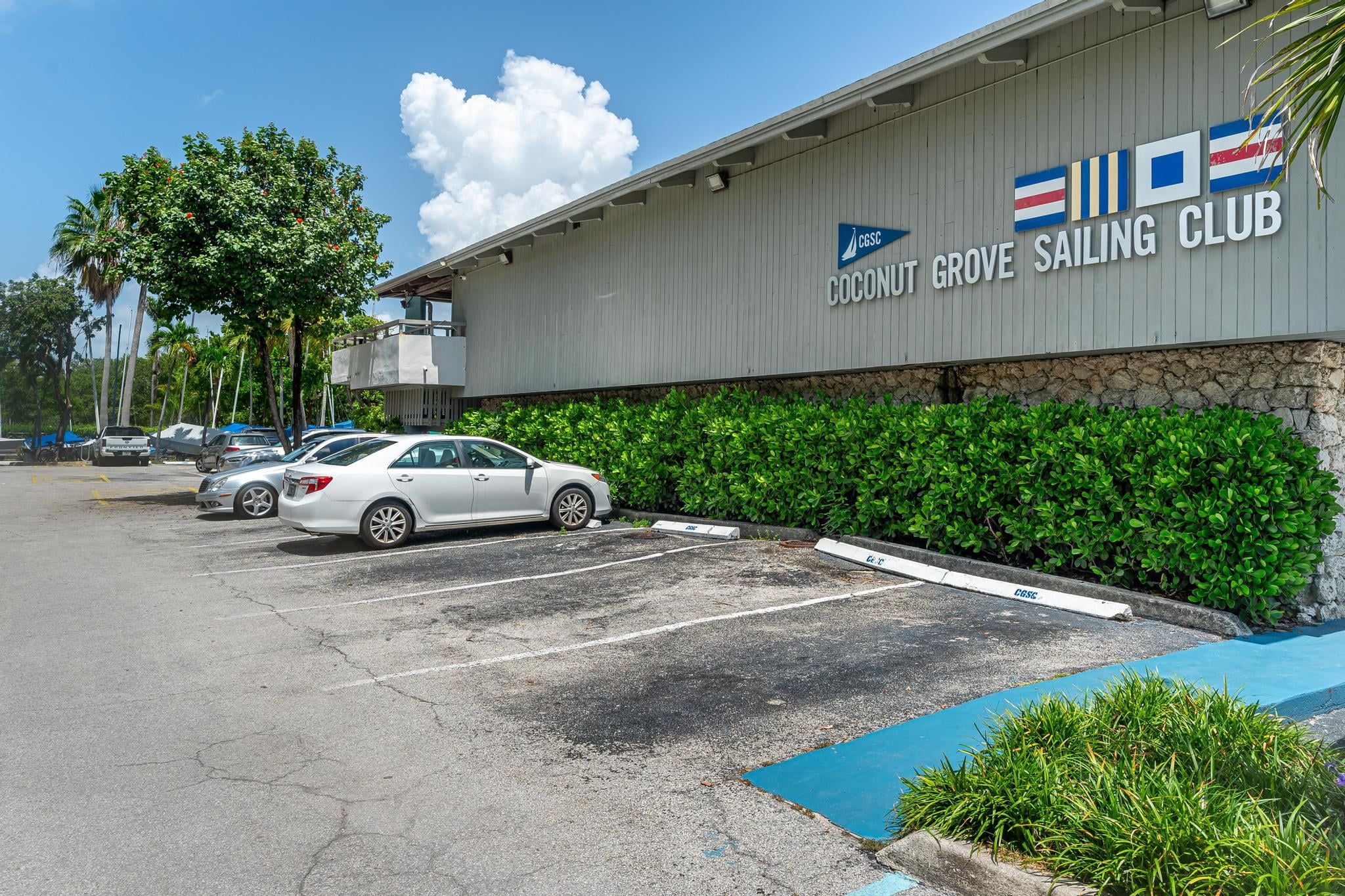 Un edificio con un letrero que indica Coconut Grove Sailing Club, espacios de estacionamiento y un cielo despejado.