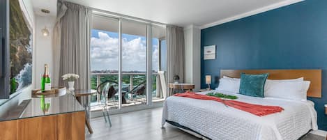 Habitación de hotel con una cama grande, un escritorio de madera, una silla, una mesita de noche y vistas a la ciudad a través de amplias ventanas.