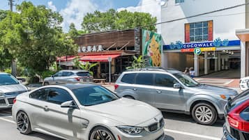 Escena callejera con automóviles estacionados, un restaurante llamado 'GUARD' y un edificio con un letrero llamativo para 'Cocowalk'.