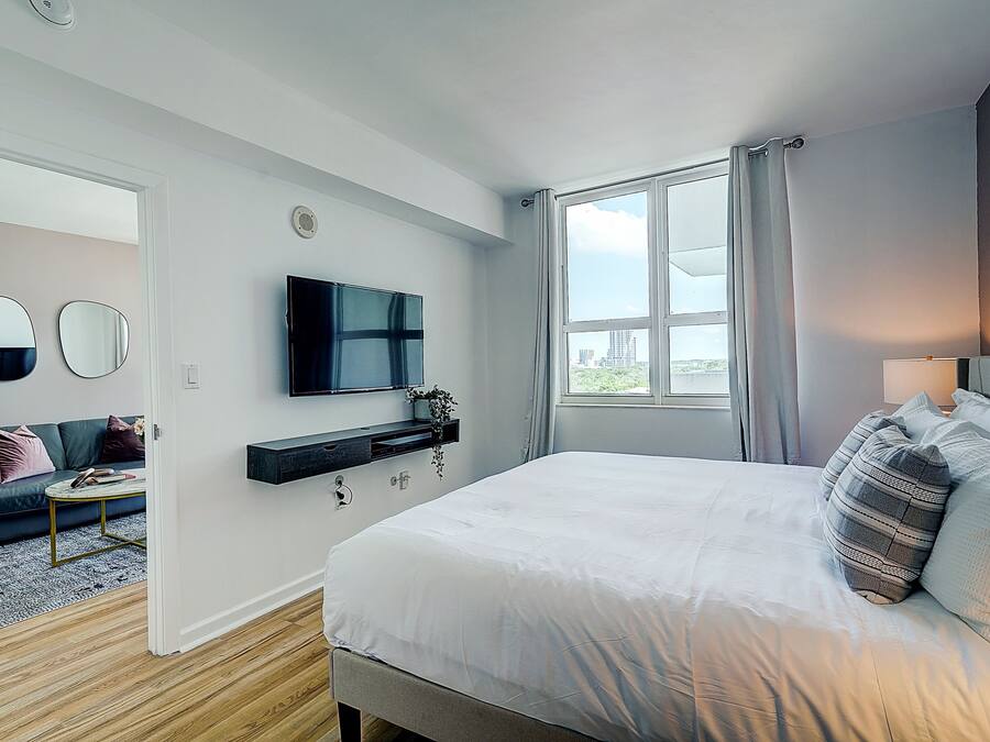 Une chambre moderne avec un grand lit, un téléviseur fixé au mur, un canapé et une fenêtre avec des rideaux.