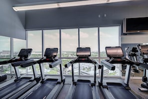 Uma academia com esteiras, televisão e grandes janelas com vista para a cidade.