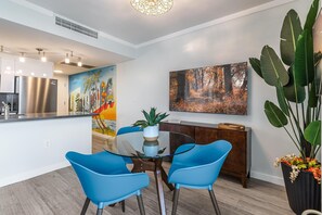 Cozinha moderna com geladeira de aço inoxidável, área de jantar com cadeiras azuis e mesa de vidro, e uma pintura grande representando uma cena de floresta.