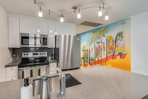 A modern kitchen with stainless steel appliances, a granite countertop, and a vibrant mural. - Bright 1BR in Coconut Grove King Bed, Views, Pool & Parking (Miami)