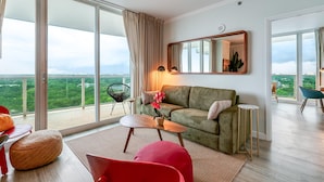 A modern living room with a green sofa, a red chair, a small table, and a large window offering a city view. - Bright 1BR in Coconut Grove King Bed, Views, Pool & Parking (Miami)