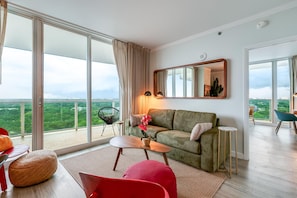A modern living room with a green sofa, a red chair, a small table, and a large window offering a city view. - Bright 1BR in Coconut Grove King Bed, Views, Pool & Parking (Miami)