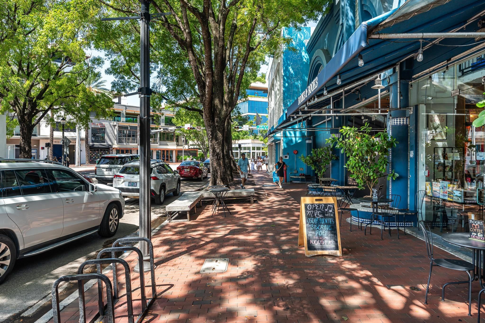Une rue bordée de voitures garées, un café avec une terrasse, et un immeuble à la façade bleue.