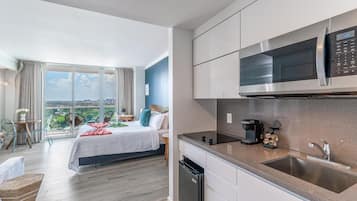 A modern hotel room with a balcony, a bed, a kitchen area with a microwave, and a coffee machine.