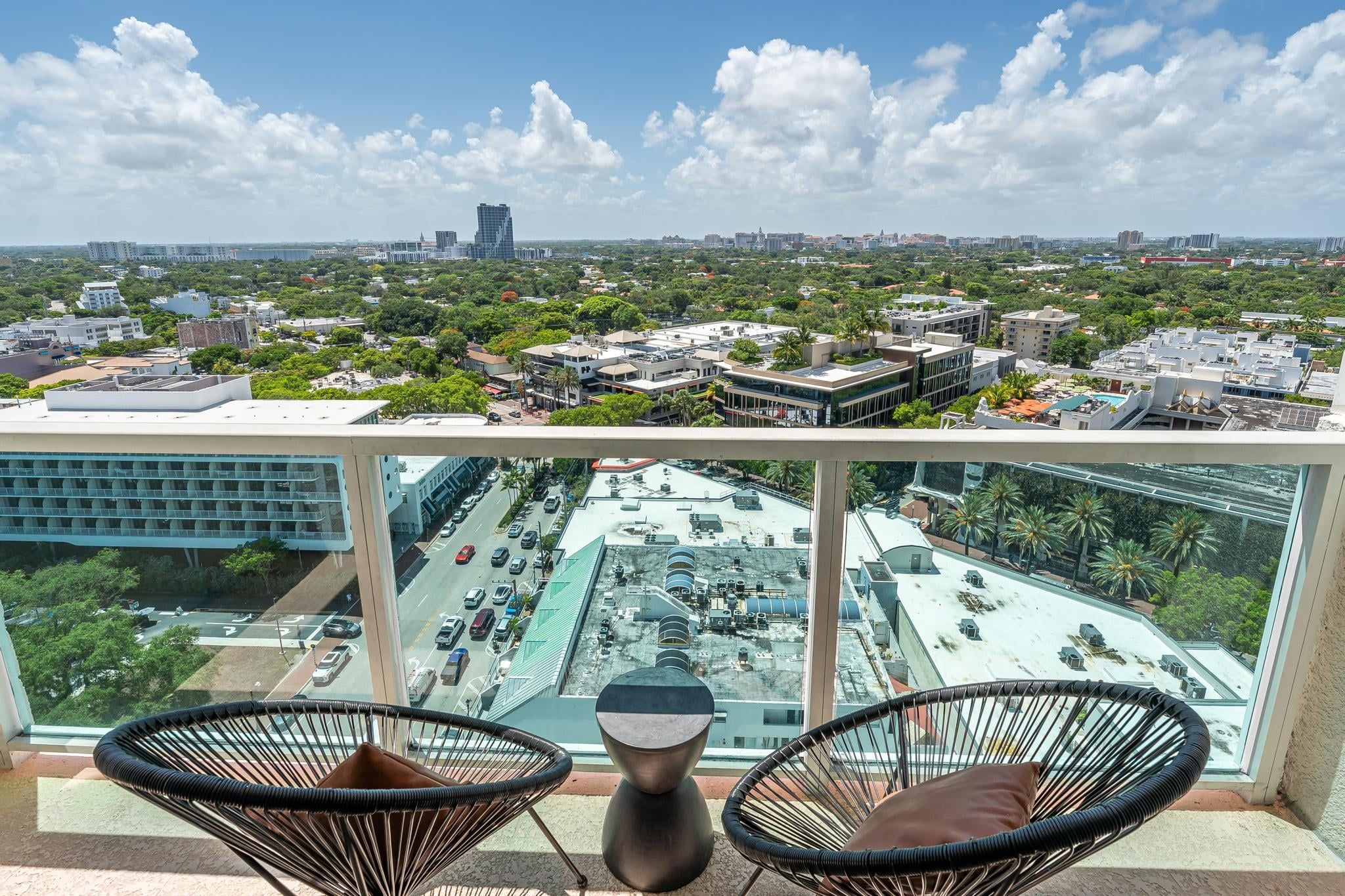 A balcony with two wicker chairs and a vase, overlooking a cityscape with buildings and a parking lot.