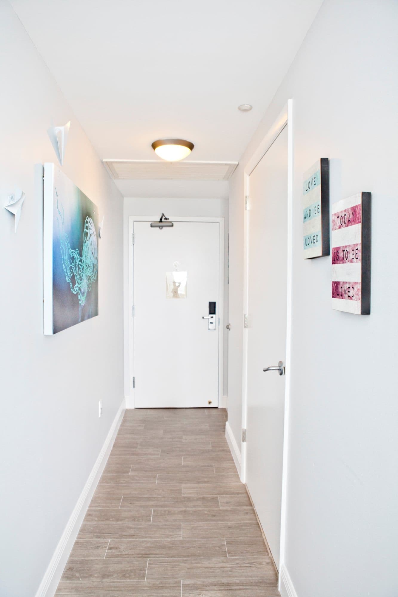 A hallway with a wooden floor, white walls, and a door at the end. There are two framed artworks on the walls and a ceiling light fixture.