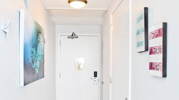 A hallway with a wooden floor, white walls, and a door at the end. There are two framed artworks on the walls and a ceiling light fixture.