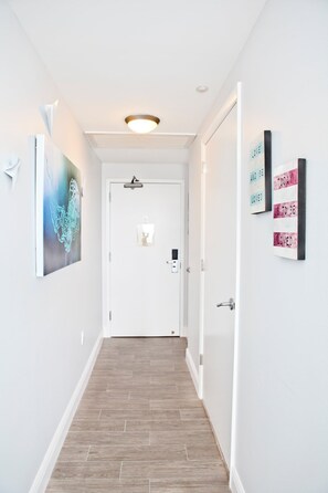 A hallway with a wooden floor, white walls, and a door at the end. There are two framed artworks on the walls and a ceiling light fixture. - Private 2/2.5 bay View Apt. in Coconut Grove Hotel Includes Parking! (Miami)