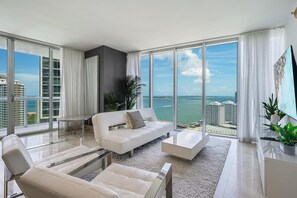 A modern living room with a white sofa, a coffee table, and a view of the ocean and cityscape through large windows. - Icon Brickell Miami- 2 Bedroom Condo With City and Water Views! (Miami)