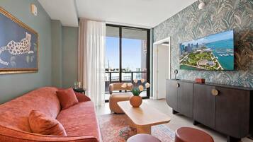 Uma sala de estar moderna com sofá, mesa de centro e apoio para os pés, uma área de jantar com mesa e uma grande janela com vista para a paisagem urbana.