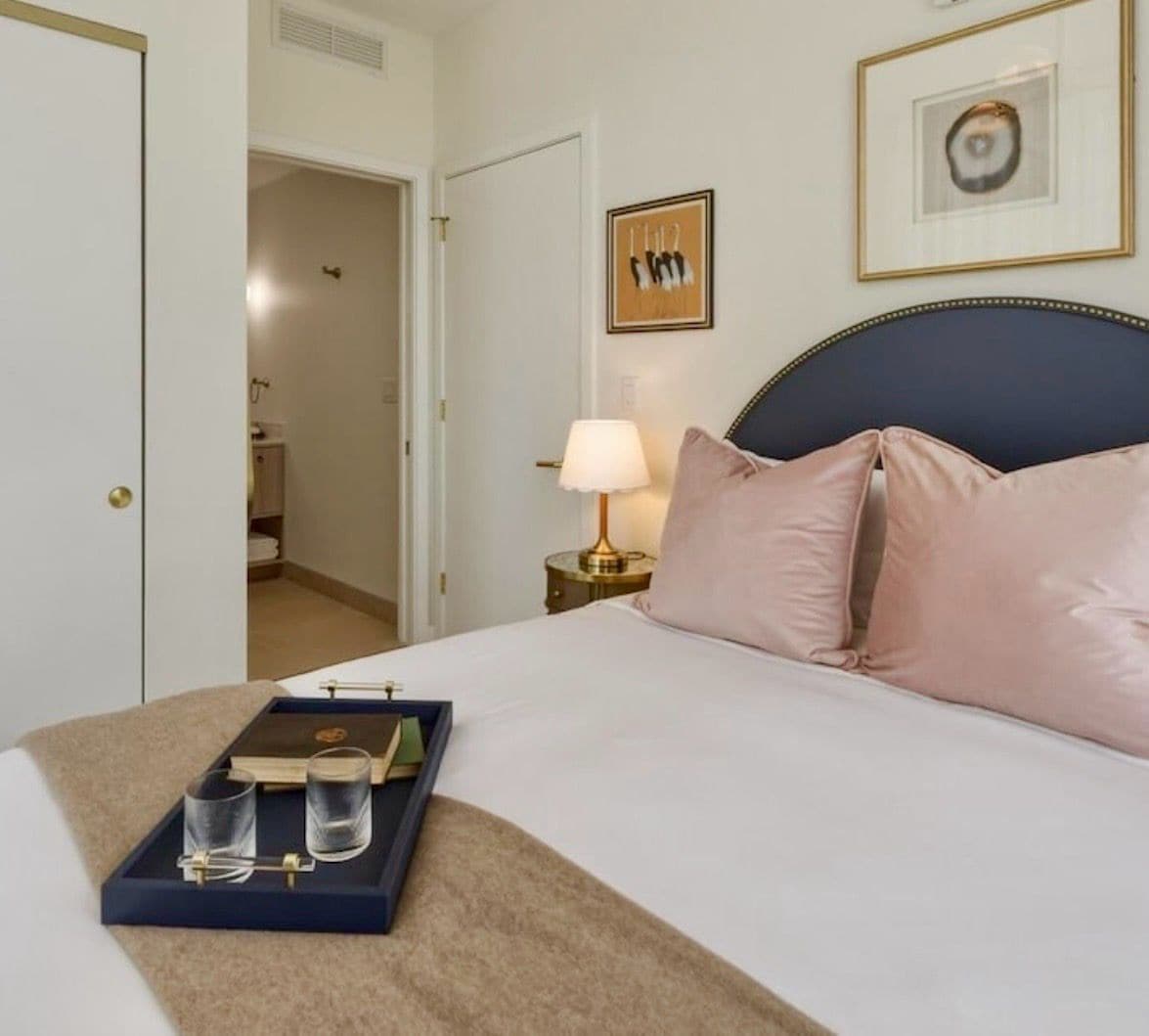 Quarto de hotel com cama, mesa de cabeceira com abajur, bandeja com copos e um livro, e uma moldura na parede.