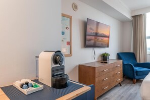 A modern hotel room with a coffee machine, a flat-screen TV, a blue armchair, and a wooden dresser. - Cozy Grove Stay Free Parking, Pool Access, Walk to Cafés & Parks (Miami)