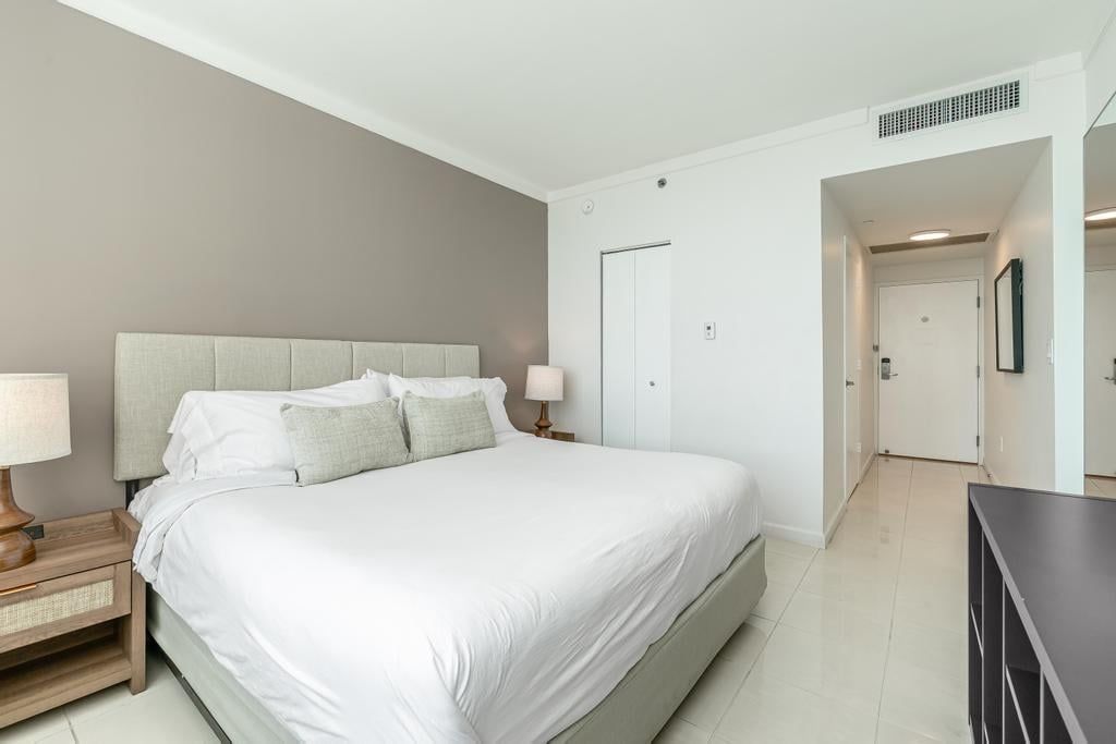 Un dormitorio con una cama, mesitas de noche, un aparador y una puerta que da a otra habitación.