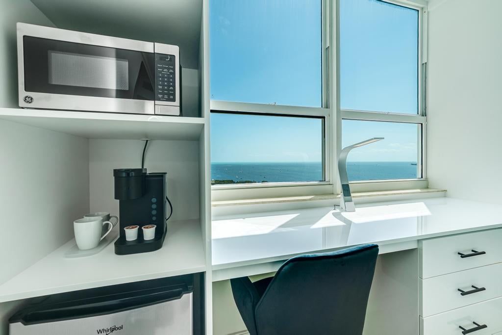 Una habitación moderna con un escritorio blanco, microondas, cafetera y vista al mar a través de un amplio ventanal.