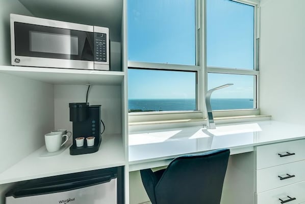 Una habitación moderna con un escritorio blanco, microondas, cafetera y vista al mar a través de un amplio ventanal.