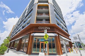 A multi-story building with a prominent 'Cucina' sign at the entrance, large glass windows, and a pedestrian crossing sign.