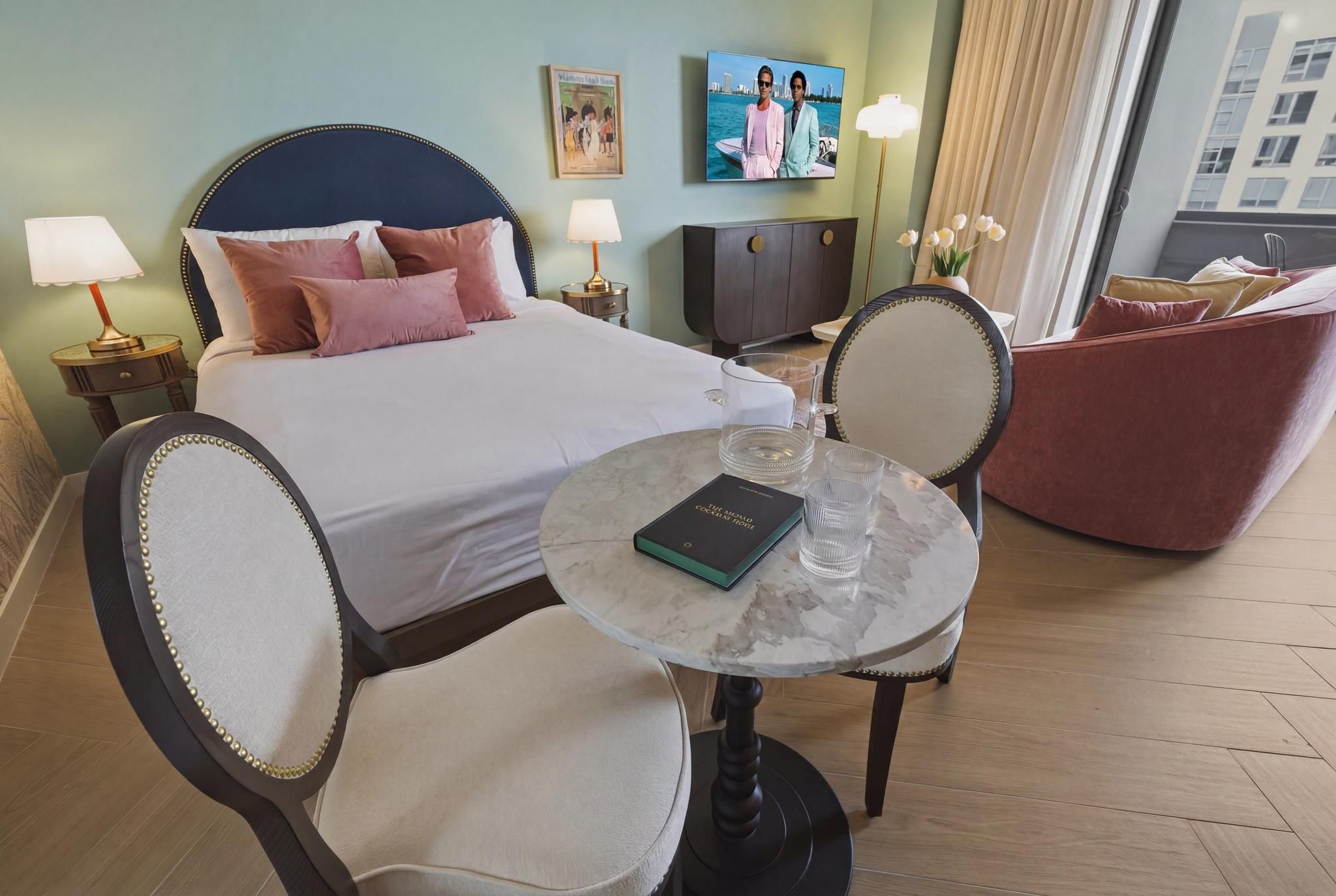 Habitación de hotel con cama, mesitas de noche, una mesa redonda con un libro, una silla, un sofá y vistas a la ciudad.