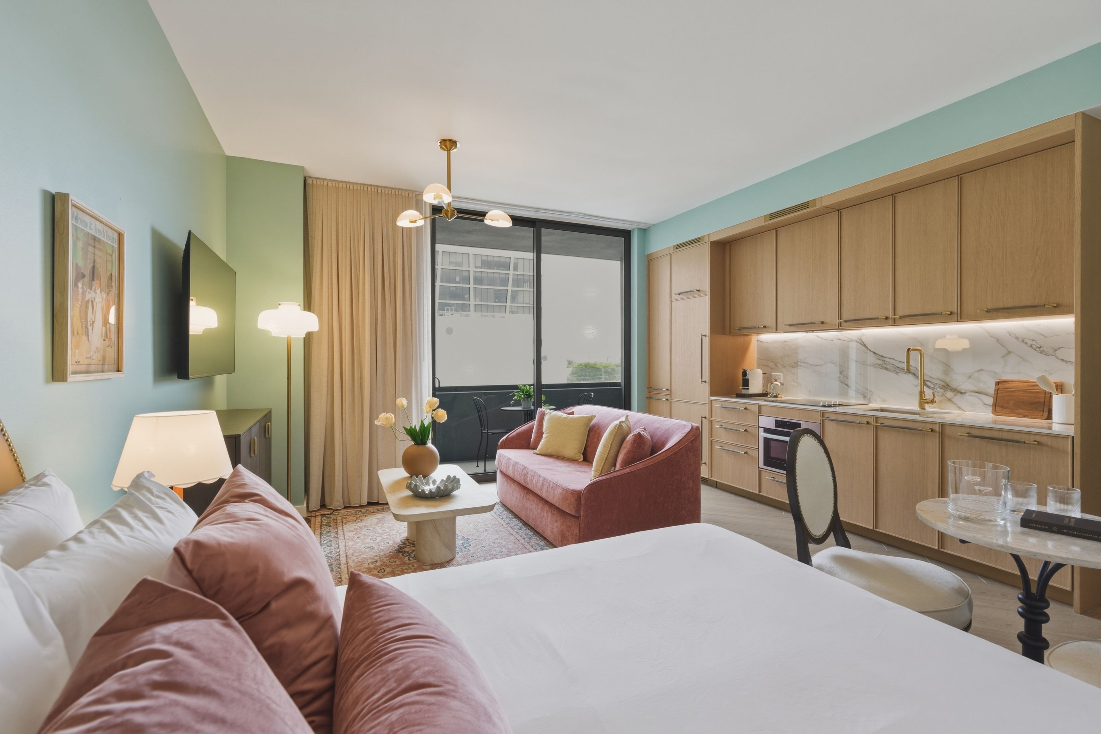 A hotel room with a bed, a sofa, a dining table, a kitchen area, and a large window.