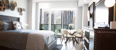 A modern hotel room with a large bed, a dining area, and a view of tall buildings.