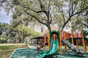 A playground with a green slide and yellow and red play structures, surrounded by trees and a building in the background. - Deluxe Studio in Coconut Grove-city & Bay Views...includes Parking (Miami)