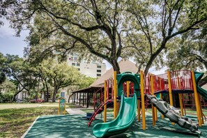 Um parquinho com um escorrega verde e estruturas de recreação amarelas e vermelhas, cercado por árvores e um edifício ao fundo.