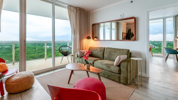 A modern living room with a green sofa, a red chair, a small table, and a large window offering a city view. - Elegant 2BR Condo at Hotel Arya Full Kitchen, Wraparound Views, Washer/dryer, 2 Parking Spots (Miami)
