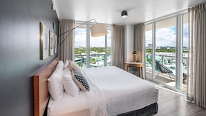 A modern hotel room with a large bed, a desk, and a view of the cityscape through floor-to-ceiling windows. - Modern Grove Studio Parking & Washer/dryer Included (Miami)