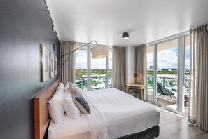 Quarto de hotel moderno com cama grande, escrivaninha e vista da cidade através de janelas do chão ao teto.