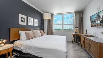 A modern hotel room with a large bed, a desk, a chair, a TV, and a window with a city view.