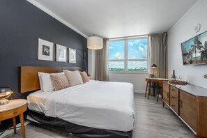 A modern hotel room with a large bed, a desk, a chair, a TV, and a window with a city view.