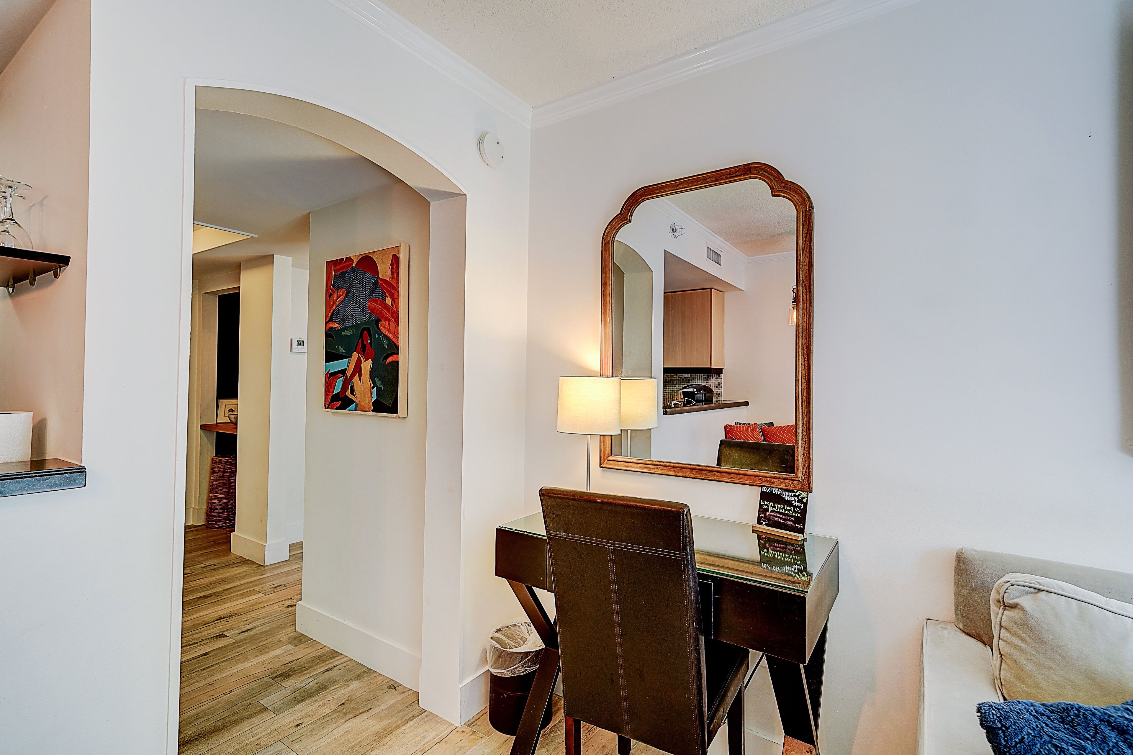 A hotel room with a desk, chair, and a large mirror. There is a lamp on the desk and a trash can next to it. The room has a wooden floor and a door leading to another room.
