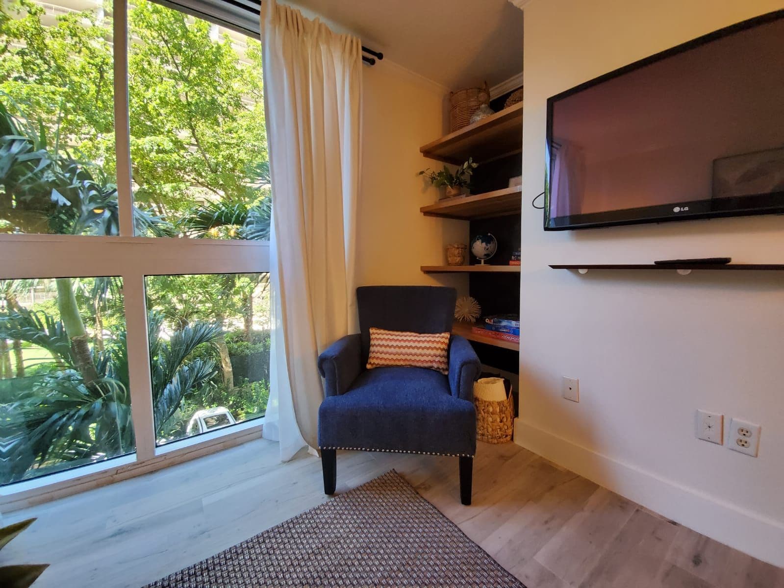 Een kamer met een groot raam, een blauwe fauteuil, een geweven vloerkleed, een plank met decoratieve objecten en een bevestigde televisie.