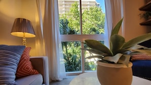 Een woonkamer met een bank, een lamp, een kamerplant en uitzicht op een gebouw en bomen door een raam.