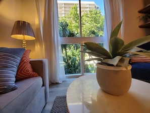 Ein Wohnzimmer mit einer Couch, einer Lampe, einem Topfpflanze und Blick auf ein Gebäude und Bäume durch ein Fenster.