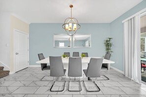 Een eethoek met een witte tafel en grijze stoelen, een kroonluchter en een grote spiegel boven de tafel.