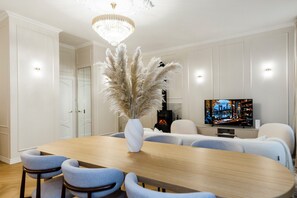A modern dining area with a large wooden table, light blue upholstered chairs, a white vase with pampas grass, a chandelier, and a television displaying a cityscape. - Elegant Apartment | Fireplace | 20 min Old Town (Riga)