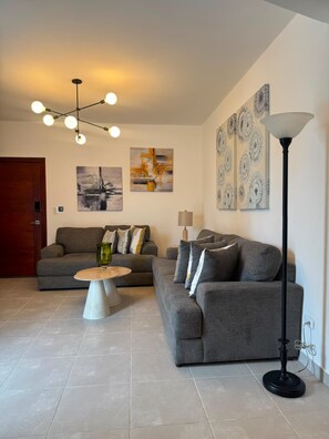 Un salon moderne avec un canapé gris, une table basse et un lampadaire. Les murs sont ornés de tableaux abstraits et une porte en bois est visible.