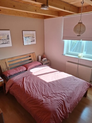 Ein Schlafzimmer mit Holzbalkendecke, einem Bett mit rosa Bettwäsche, einem Nachttisch mit Lampe und zwei gerahmten Bildern an der Wand.