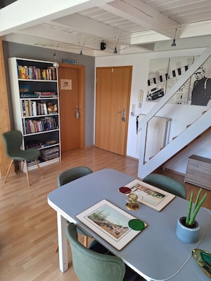 Ein Zimmer mit Esstisch, Stühlen, einem Bücherregal, einer Treppe und einer Tür mit der Aufschrift 'STRAND'.