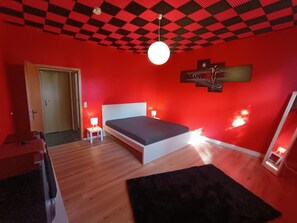 A room with a bed, a small table with a lamp, and a red wall. - Vrbo Property (Joachimsthal)